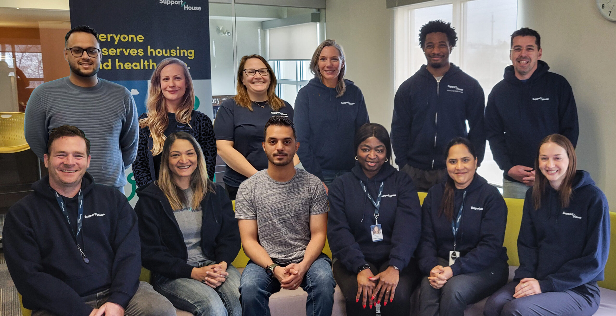 A group of thirteen people are posed together indoors for a team photo. Five individuals are seated in the front row and eight are standing behind them. Most are wearing dark hooded sweatshirts with a small logo on the chest. They are in a bright office space with windows, chairs, and a banner in the background that reads ‘Everyone deserves housing and health.