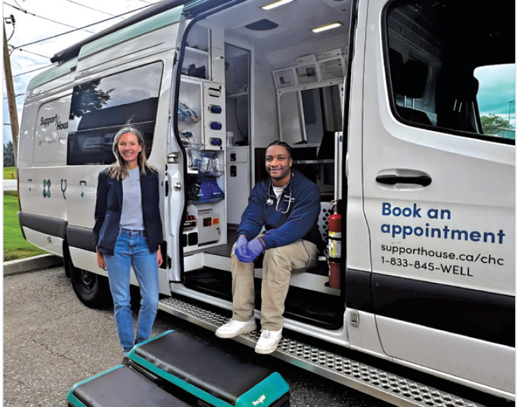 Two individuals stand beside an open mobile health van; one individual is standing on the pavement near the doorway, while another is seated on the van’s interior step, with medical equipment visible inside and signage on the van promoting appointment booking.
