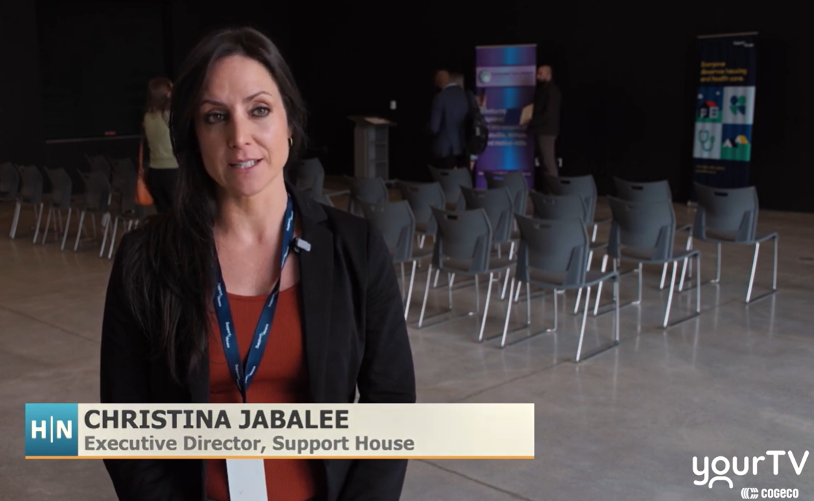 Person speaking on camera during a news interview at an indoor event space, with rows of chairs, banners, and a stage visible in the background. On-screen text reads: “Christina Jabalee, Executive Director, Support House,” with a YourTV logo.